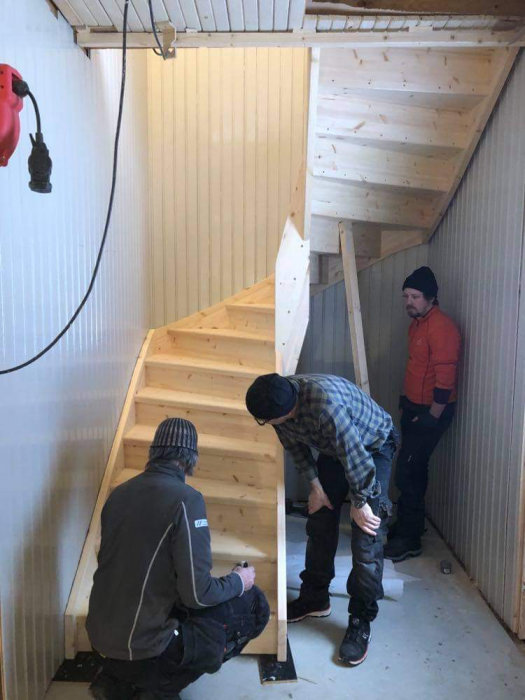 Två personer monterar en trätrappa inomhus med ojämna väggar och tak.