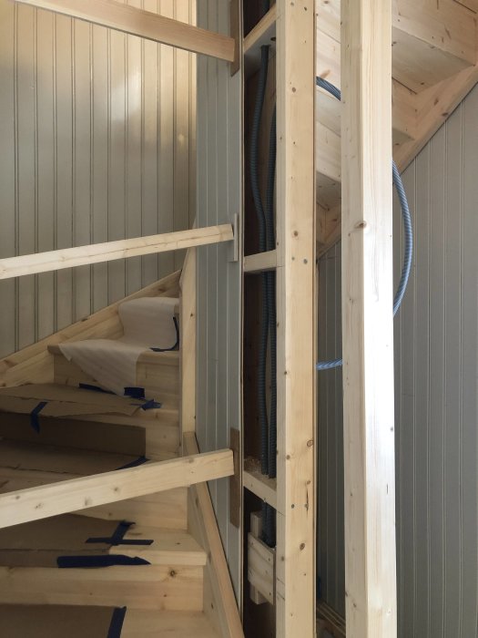 Hörn av ett trapphus under konstruktion med trätrappor och ställning, tejpade steg och väggrör.