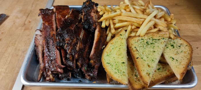 Grillade revbensspjäll med pommes frites och vitlöksbröd på en serveringsbricka.