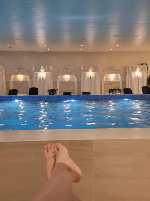 Inomhuspool med eleganta vita pelare och speglar, betraktad från kanten med synliga fötter.