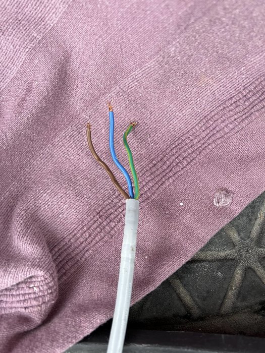 Elektrisk kabel med avskalade brun, blå och grön/gul trådar mot en lila textilbakgrund.
