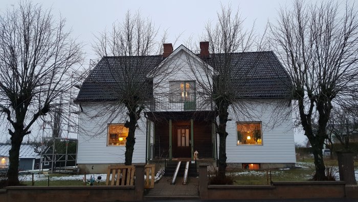 Ett tvåvåningshus med vit PVC-fasad, upplyst fönster, snöfläckad gräsmatta och nakna träd.