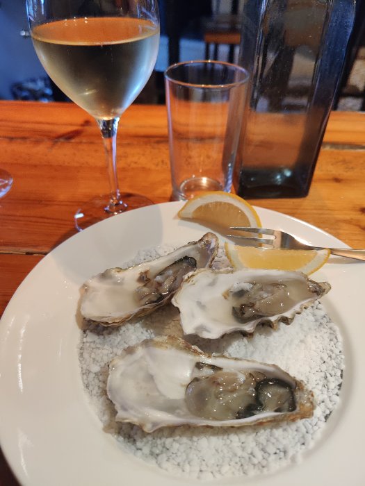 Tallrik med ostron, citronskiva, och glas med vitt vin på ett träbord.