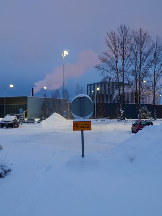Parkering med skylt som säger "Camping förbjuden" i snöigt landskap med industri och gatubelysning i skymning.
