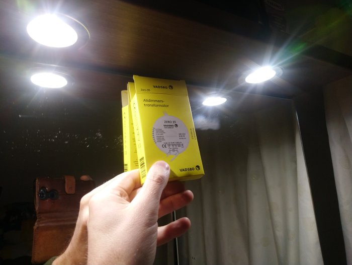 Hand håller en förpackning för en Vadsbo transformator med LED-taklampor tända i bakgrunden.