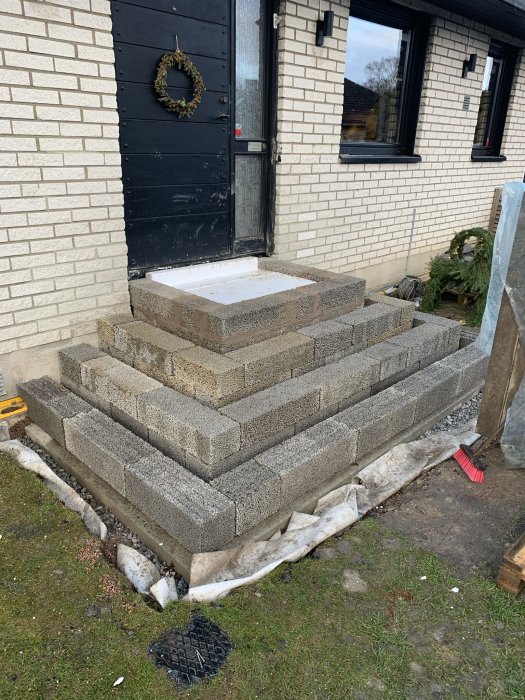 Obyggd stentrappa med flera steg av lecablock framför en husentré, med byggmaterial och en röd sopborste synlig.