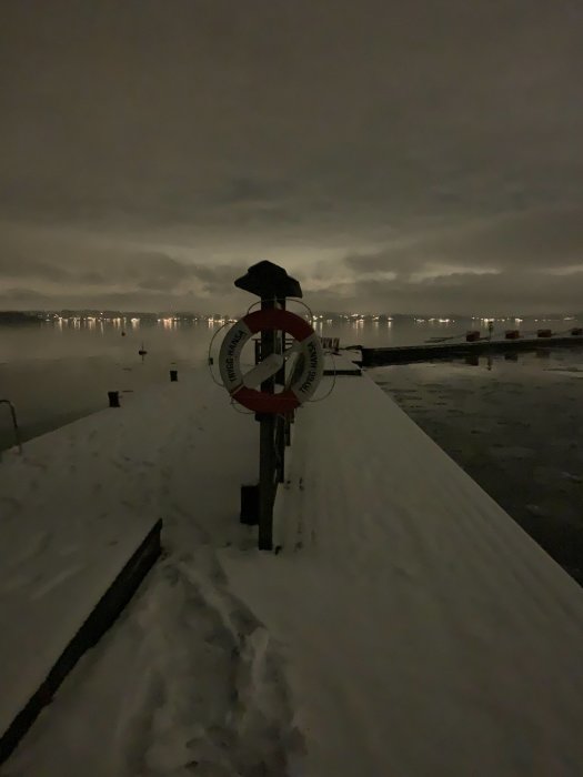 Mörk vinterbild på en brygga med snö, livboj upphängd på stolpe och ljus från en stad i bakgrunden.