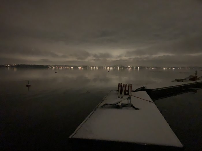 Nattbild över sjö med brygga täckt av snö och ljus från bebyggelse i bakgrunden.