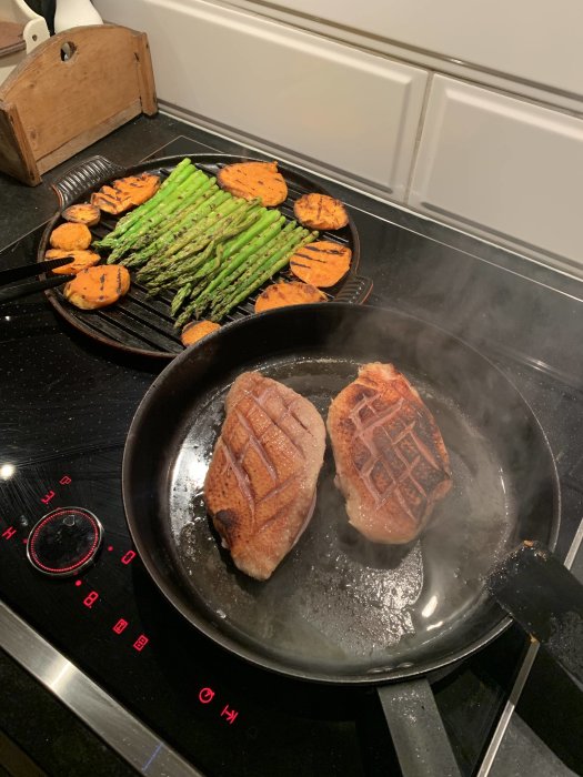 Anka tillagas i stekpanna, med sparris och sötpotatis på grillpanna över spishäll.