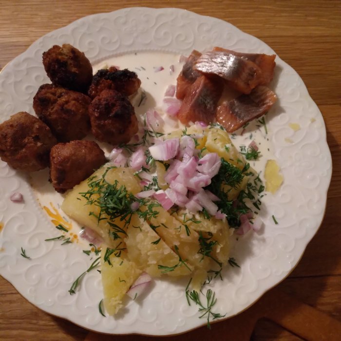 Midsommartallrik med sill, potatis, köttbullar, hackad rödlök och dill på ett vitt mönstrat fat.