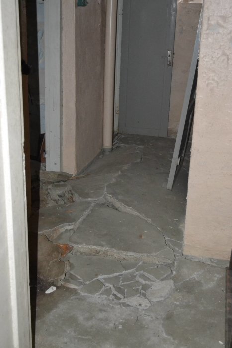 Sönderfrusen betongplatta i källare med sprickbildning och avbrutna bitar vid en väggkant.
