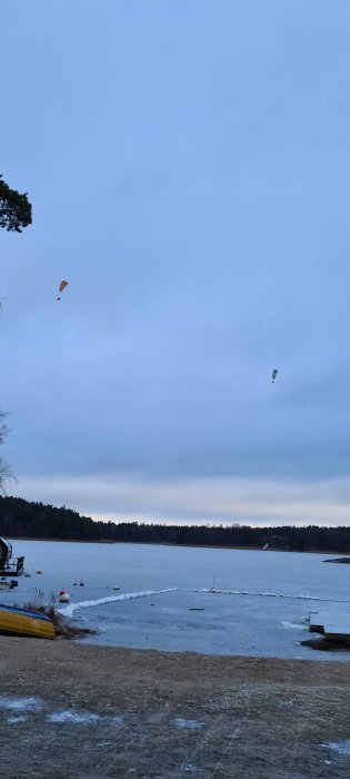 En frusen sjö vid skymning med tre luftburna drakar och en gul kanot på stranden.