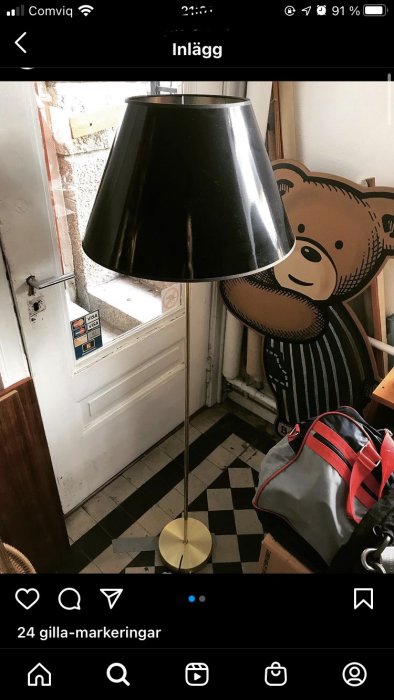 Golvstående lampa med svart skärm och guldig fot framför en dörr med ett nallebjörnsmotiv, indikerar fråga om elektrisk säkerhet.