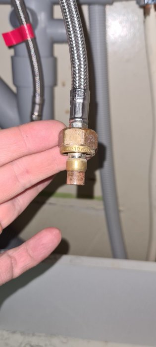 Hand håller en vattenanslutningsdetalj märkt med "10", som syns framför rörinstallationer.