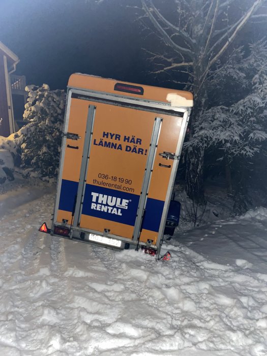 Bakifrån bild av ett hyrsläp i snön, högerbakhjul fast i ett dike och släpvagnen nuddar marken.