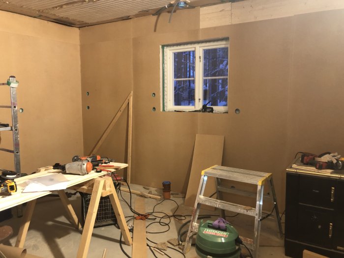 Renoveringsarbete med nya masonitskivor på vägg i ett rum med verktyg och byggmaterial.