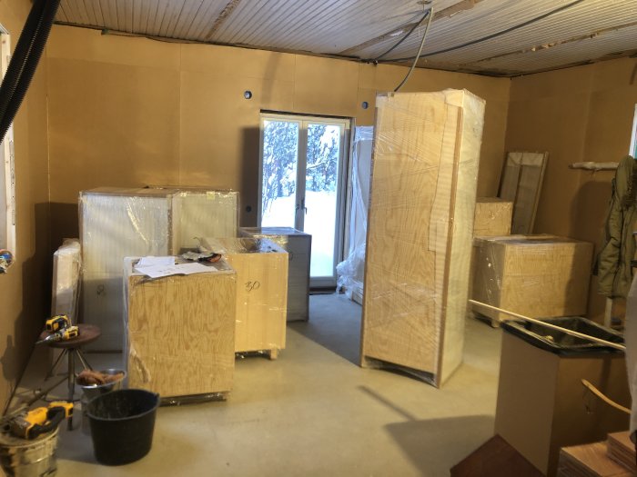 Renoveringsrum med inplastade köksmöbler, verktyg, och masonitbeklädda väggar.
