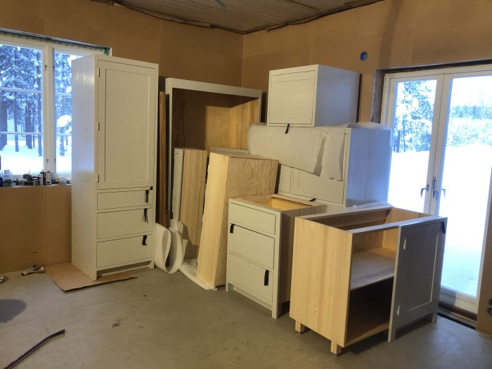 Opackade köksskåp och lådor staplade i ett rum under renovering med snö synlig genom fönstren.