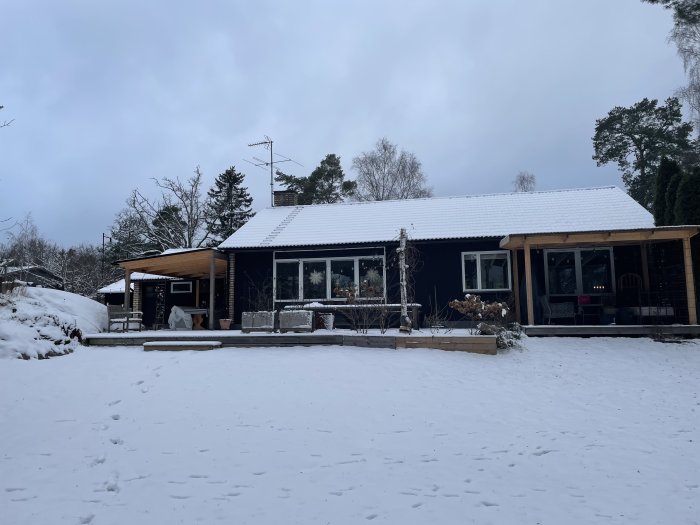 Ett enplanshus med snötäckt tak och en altan i ett vinterlandskap, synliga fotspår i snön framför huset.