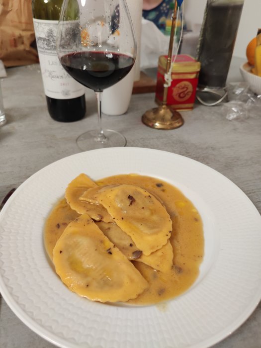 Ett fat med tryffelravioli i en krämig sås, bredvid ett glas rött vin och en vinflaska i bakgrunden.