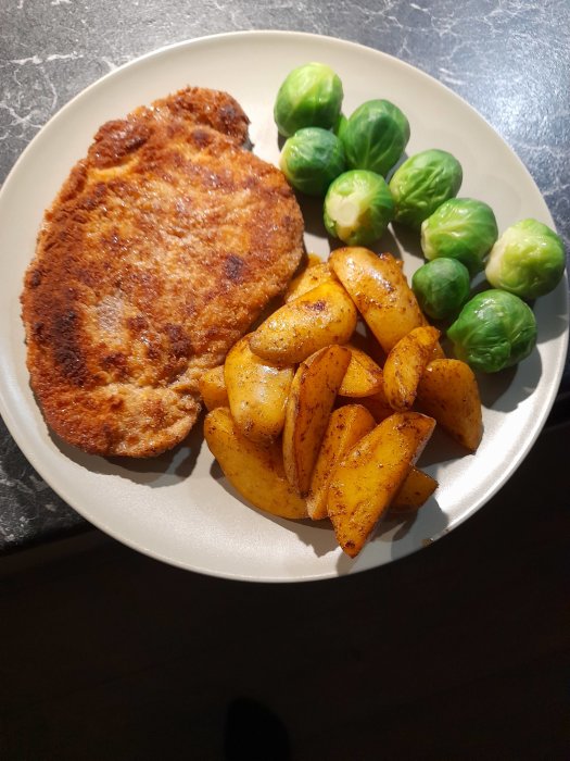 Stekt schnitzel med kryddade potatisklyftor och brysselkål på en tallrik.