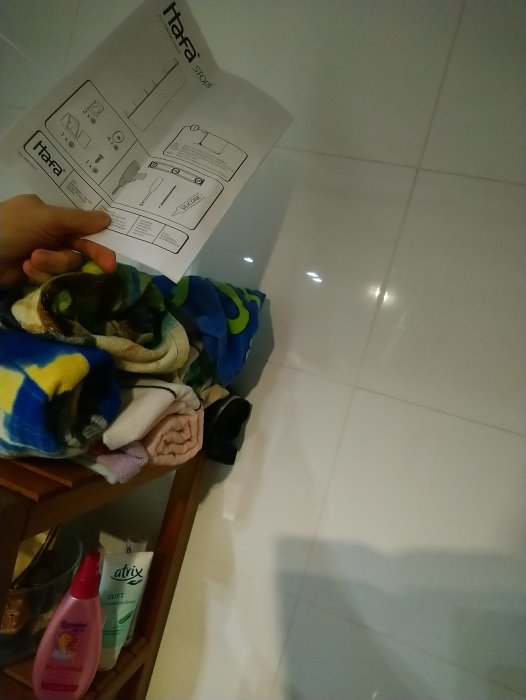 Anvisning för badrumsskåp hålls upp framför kaklade vägg, trångt badrum med handdukar och hygienprodukter synliga.