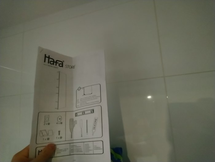 Hand håller upp en instruktionsmanual för badrumsskåp framför vit kaklad vägg.