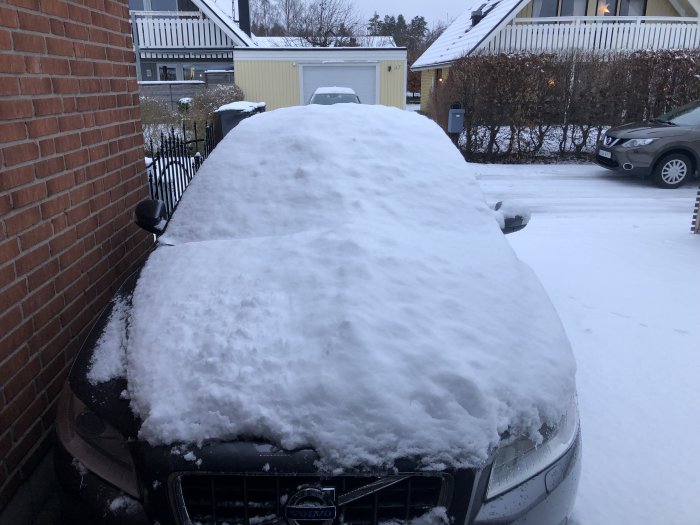 Bil täckt av tjockt lager snö parkerad vid hus på vinterdag.