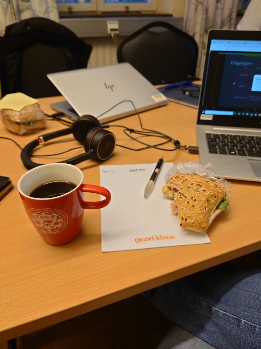 Arbetsplats med öppen laptop, hörlurar, smörgås och kaffe. Texten "good ideas" syns på ett papper.