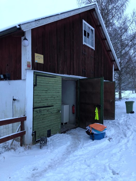 Vinterbild på ett öppet garage med rödväggade väggar, gröna portar och snötäckt mark.