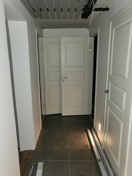 Korridor i ett hus med synliga 22mm kopparledningar i taket, grått klinkergolv och vita dörrar.