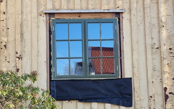 Nyinstallerat fönster på en fasad med gulnande träväggar och en reflekterande takkupa i rutan.