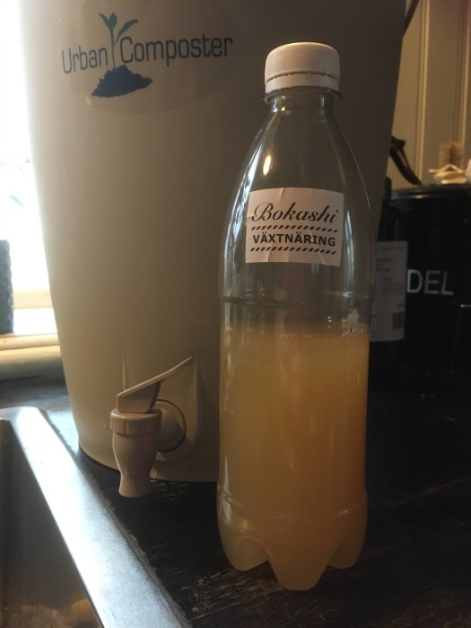 En petflaska märkt "Bokashi växtnäring" fylld med lakvatten bredvid en Urban Composter.