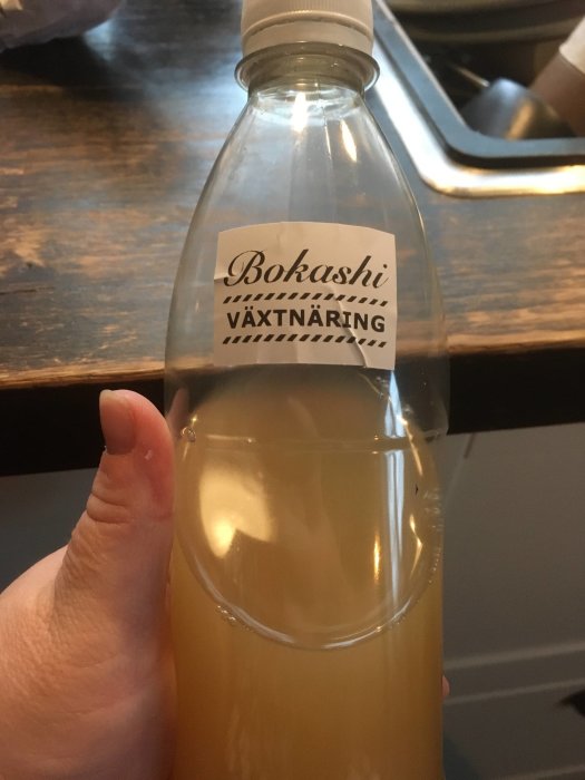 En hand som håller en klar petflaska med etiketten "Bokashi VÄXTNÄRING" fylld till hälften med gulaktigt lakvatten.