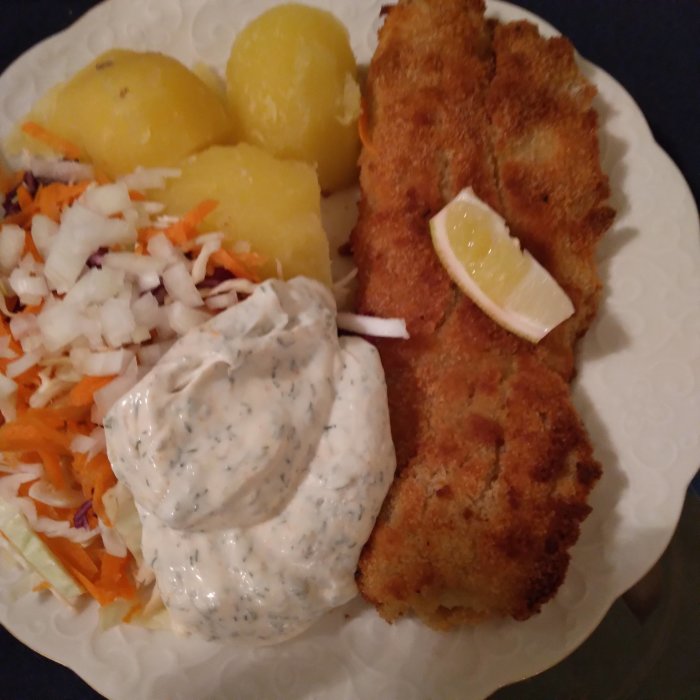 Panerad fisk med dillmajonnäs, kokt potatis, sallad och citron på en vit tallrik.