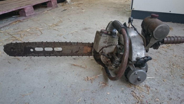 Gammal sliten motorsåg med rostspräcklor och sliten kedja ligger på ett betonggolv med sågspån.