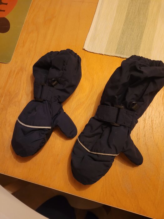 Ett par svarta vinterhandskar med spännen ligger på ett ljust trämöblerat underlag.