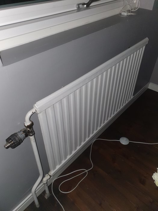 Väggmonterad radiator under fönster med synliga rör och termostat, på ett mörkt trägolv.