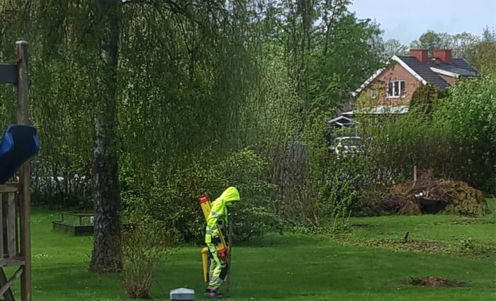 En person i hög synlighetsklädsel mäter tomtgräns med en totalstation i en grön trädgård.