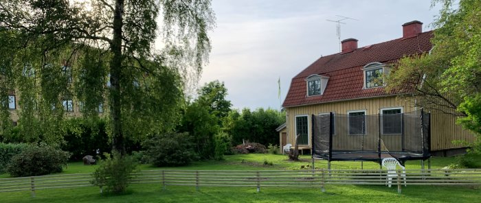 Trädgård med staket framför en gul villa, svenska flaggan och en studsmatta.