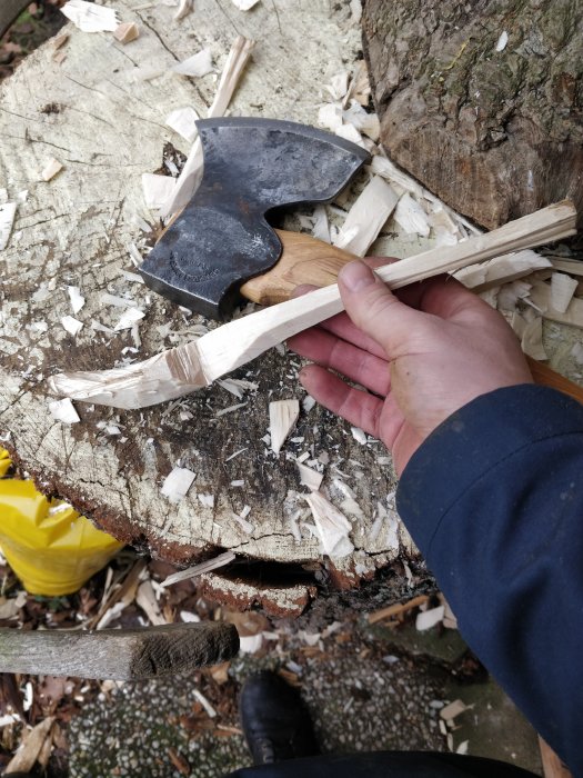 Yxa i hånden splitter ved på en trästubbe, med utspridda träflis och otydliga föremål i bakgrunden.