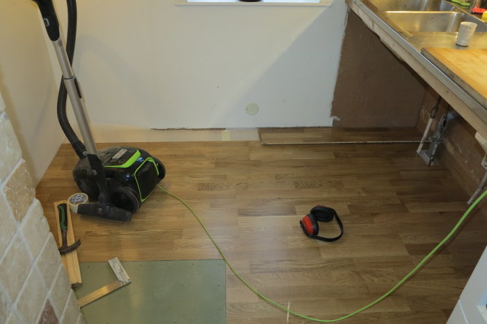 Parkettgolv nästan färdiglagt i kök med verktyg och dammsugare; en mindre del vid väggen är ofullständig.
