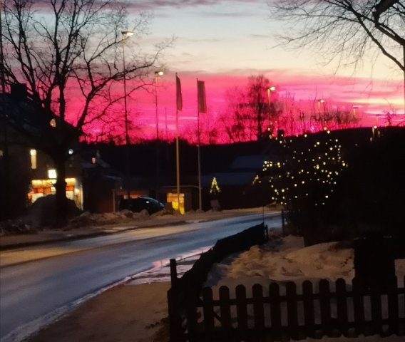 Vintergata vid skymningen med rosa himmel, silhuetter av träd och ljusslinga längs staketet.