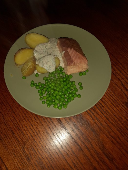 Tallrik med hemmarökt lax, kokt potatis, ärter och dillsås på ett träbord.