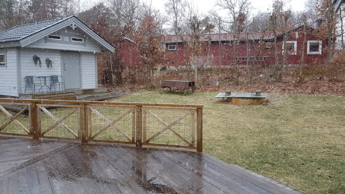 Våt trädäck med fiskbensmönster nära en stuga under snöfall, med grå himmel och trädgård i bakgrunden.