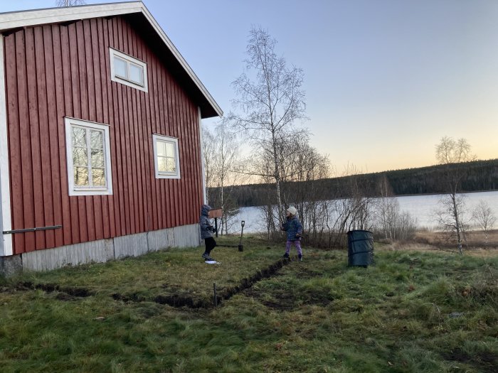 Plats där altan planeras framför röd stuga med två personer som gräver, sjö i bakgrunden.