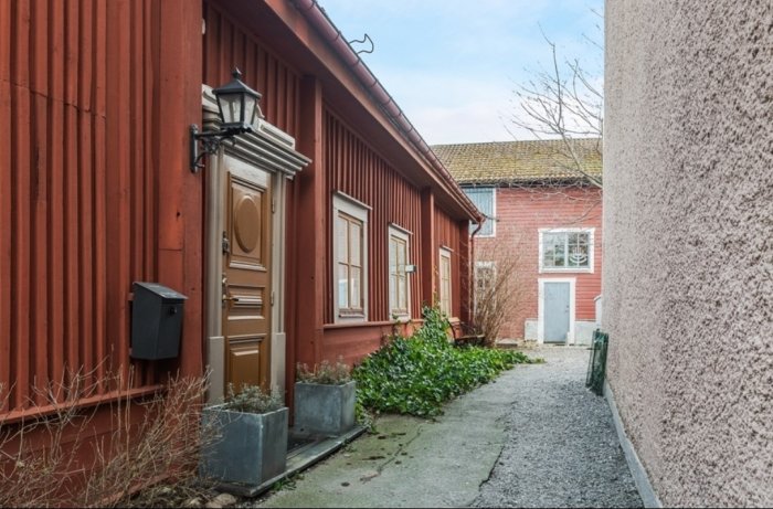 Smal gränd med rödfärgade trähus och kullerstenar, leder mot en innergård i historisk miljö.