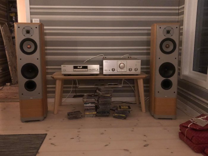 Två golvstående högtalare med en förstärkare och CD-spelare mellan sig på ett litet bord, omgivna av CD-skivor.
