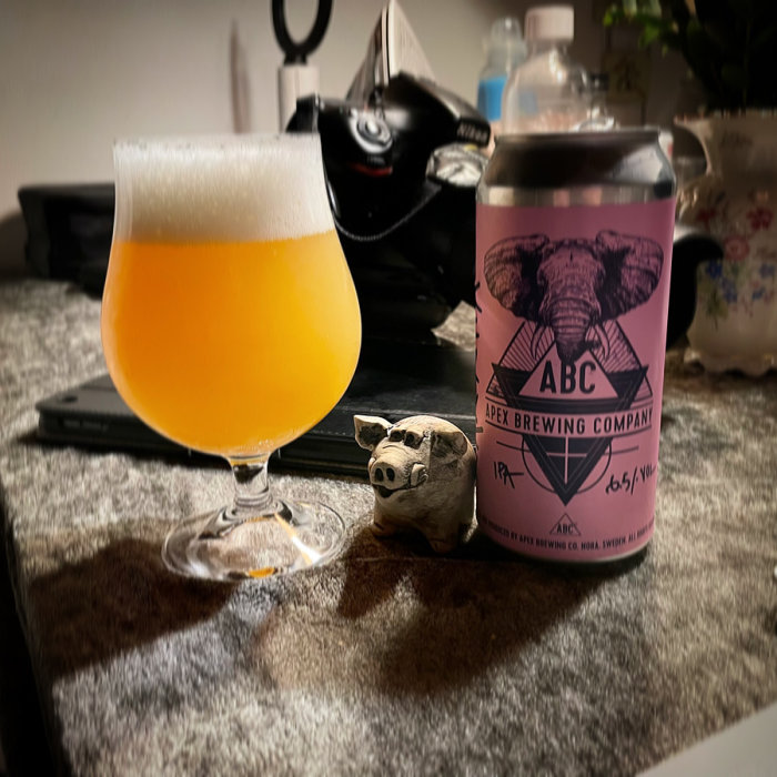 Apex Brewing Company ölburk och ett glas öl på bord med prydnadsgris och suddiga bakgrundsobjekt.