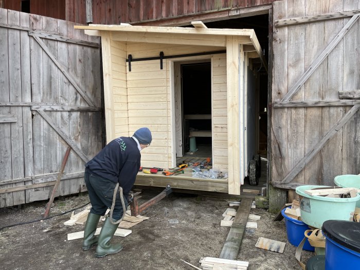 Person bygger bastu av ljust trä på en gammal balkärra bredvid ett mörkt träskjul.